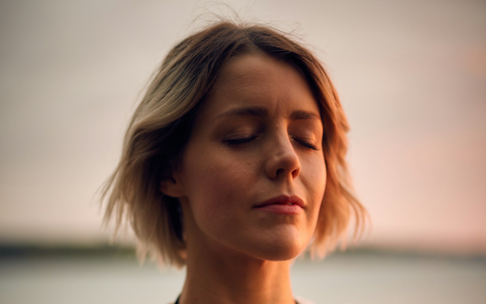 Woman with eyes closed breathing calmly by the water, her face bathed in warm golden evening light.