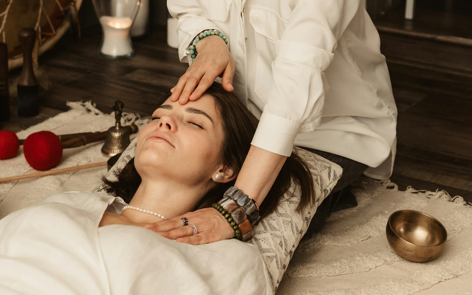 Practitioner dressed in white gently resting her hands on the forehead of a relaxed woman lying down, a candle and singing bowl nearby.